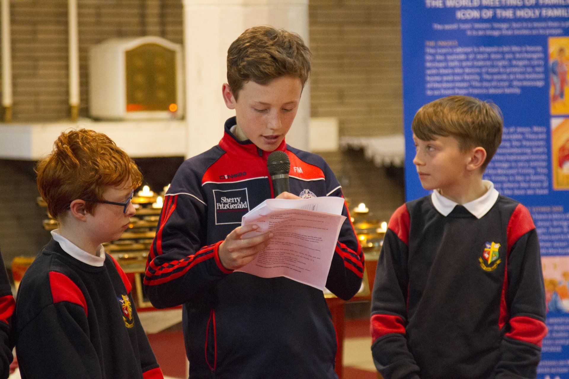 A boy is holding a microphone and reading a piece of paper while two other boys look on.