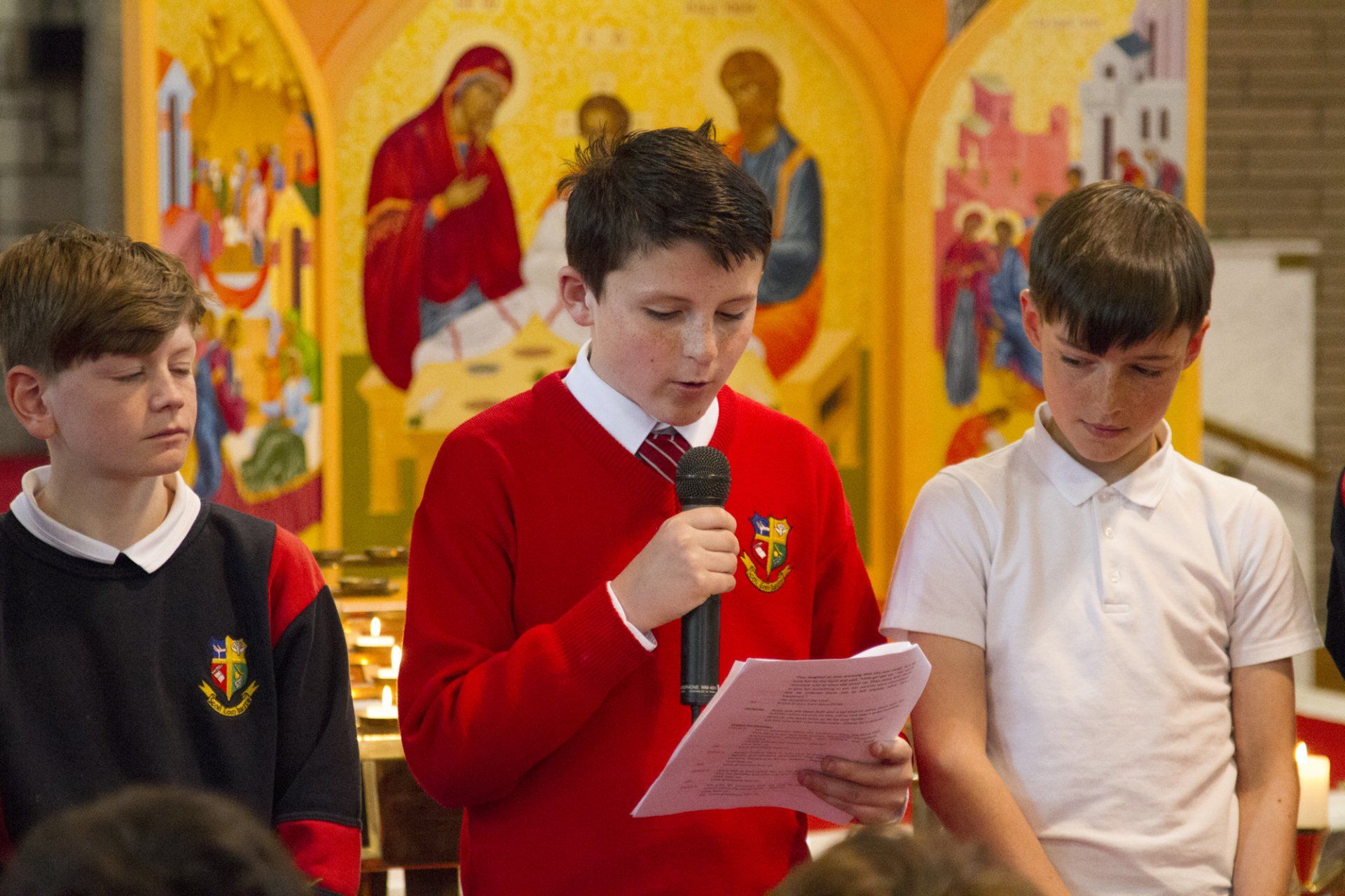 A boy in a red sweater is holding a microphone and reading a piece of paper.