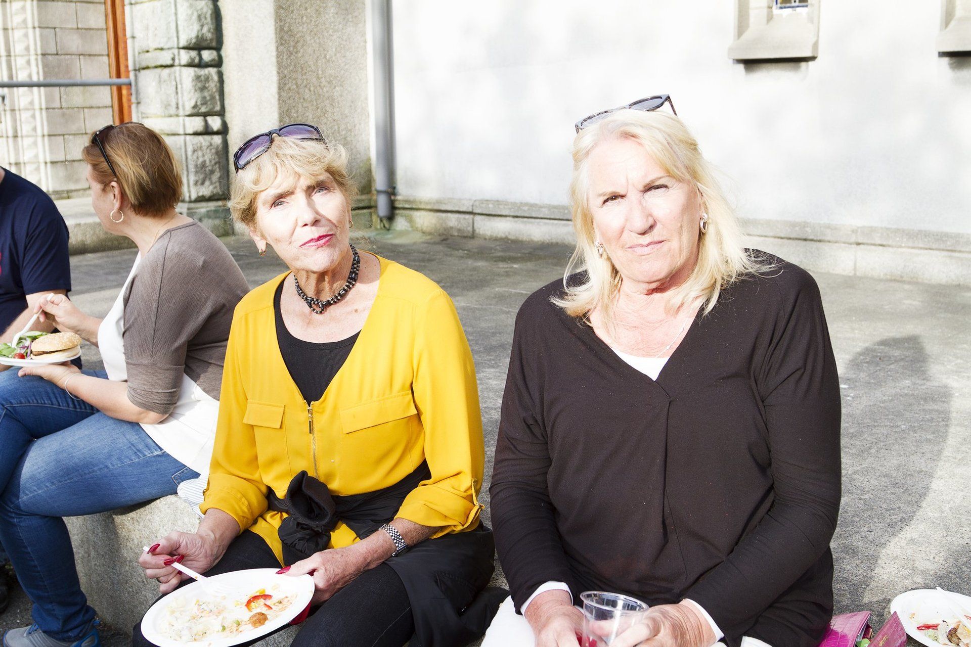 Two women are sitting on a bench with plates of food in their hands.
