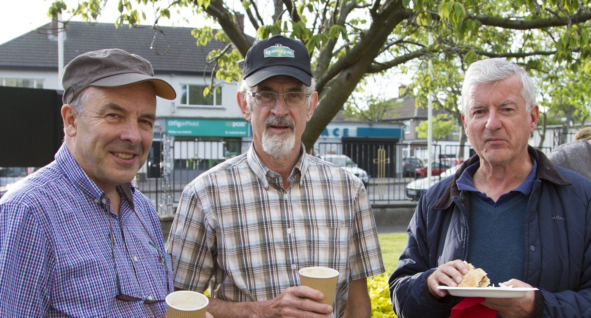 Three men are standing next to each other holding cups of coffee and a plate of food.