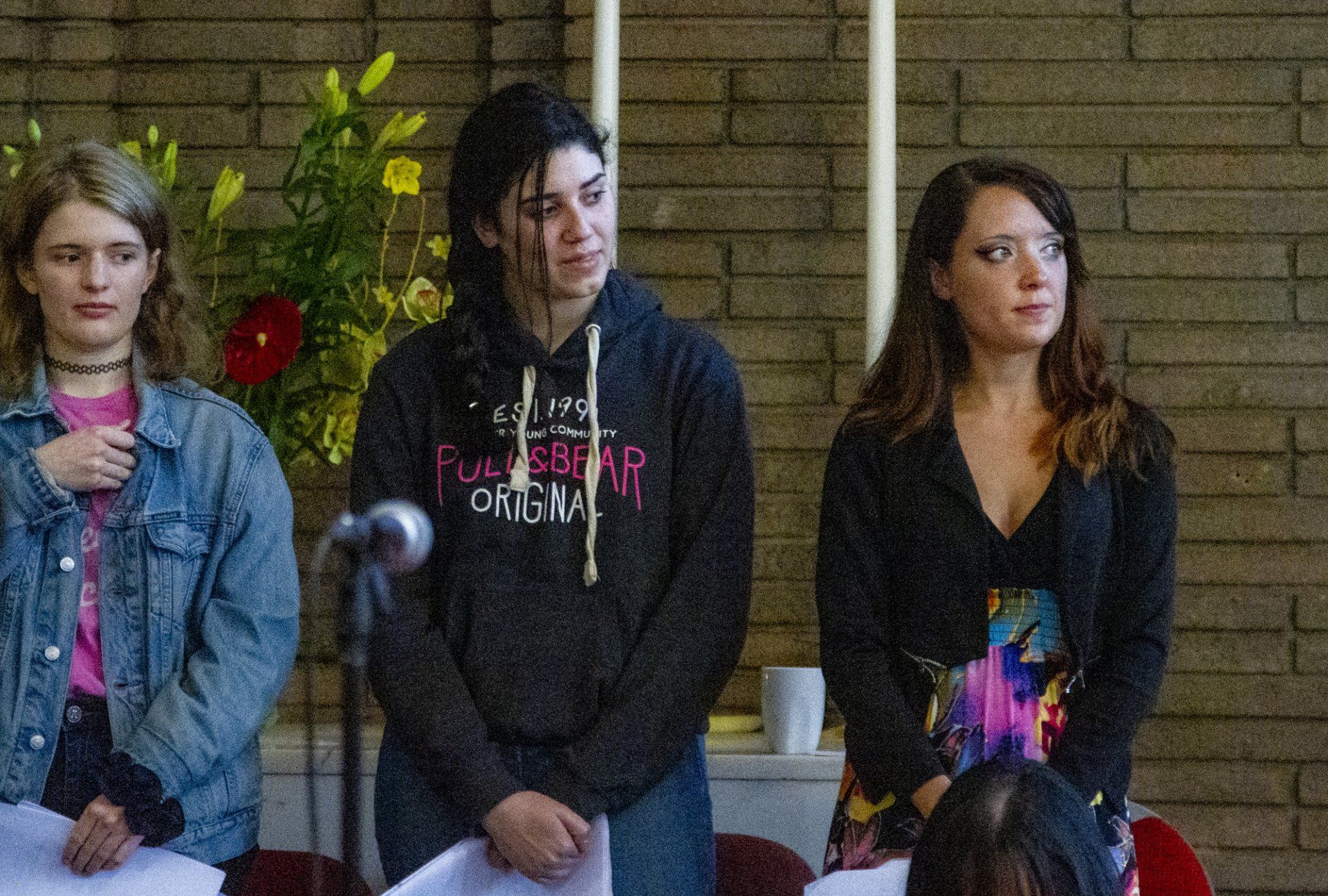 Three women are standing in front of a microphone in front of a brick wall.