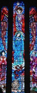 A stained glass window in a church shows a woman surrounded by angels and a lion.