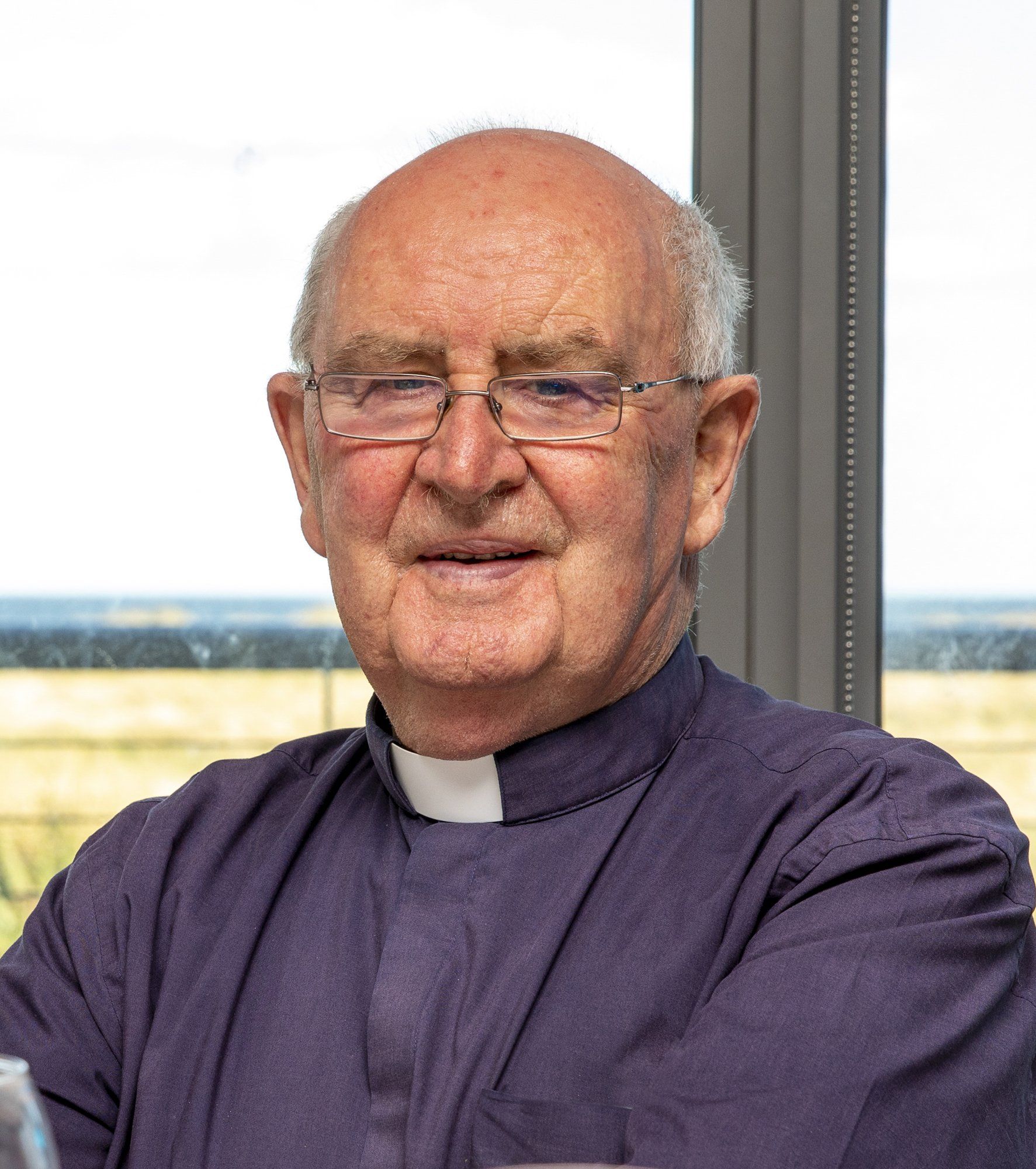 A man wearing glasses and a purple shirt is standing in front of a window.