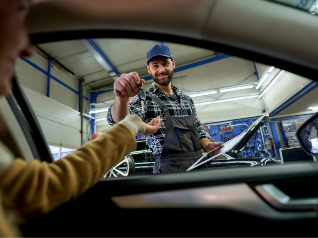 A smiling mechanic in a workshop hands car keys to a customer sitting inside a vehicle. | Russell's Garage