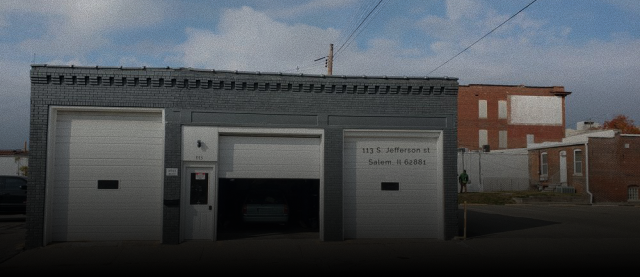 A gray, one-story garage with three white bay doors, one of which is open, set against a blue, cloudy sky. | Russell's Garage