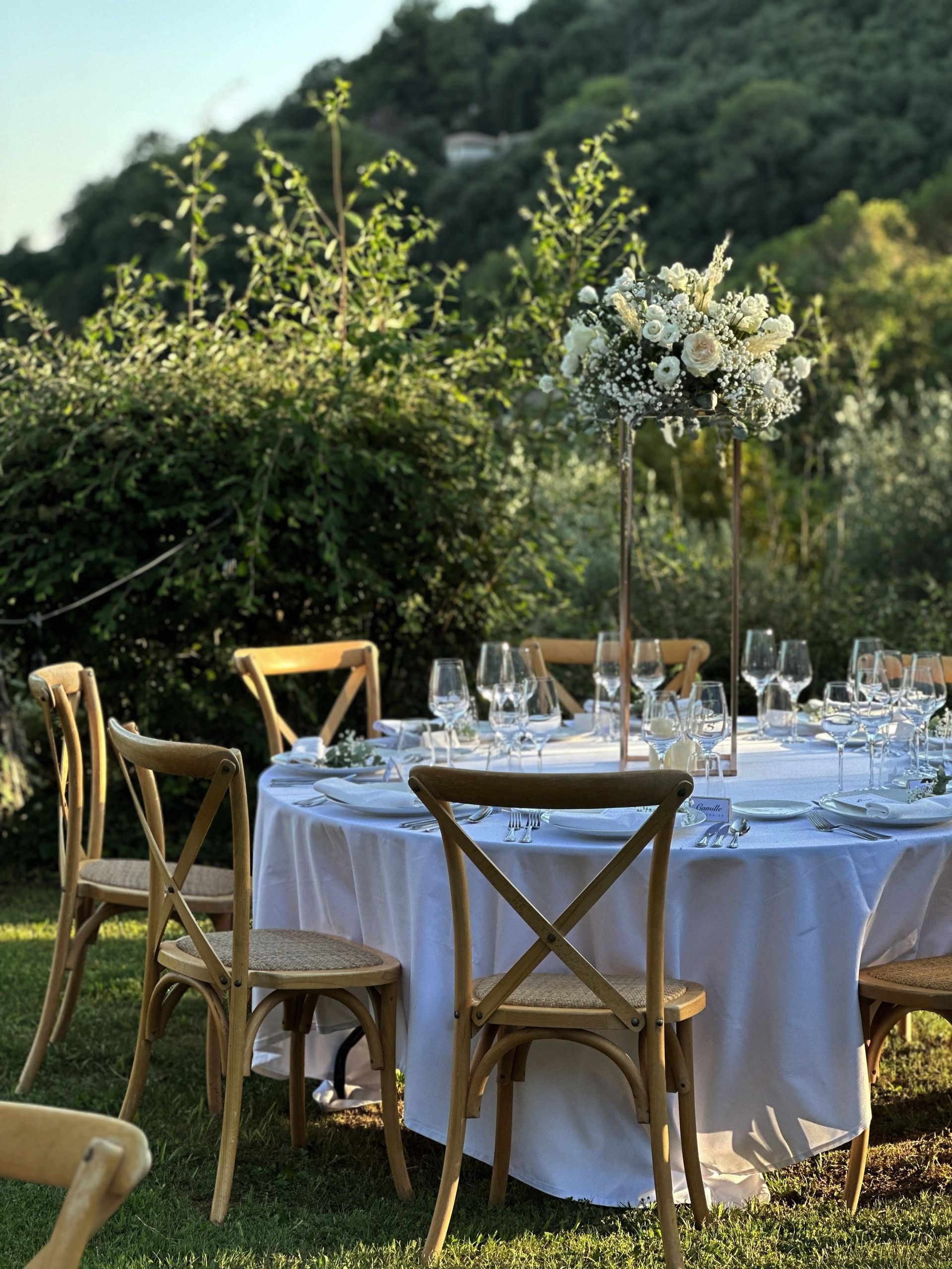 A table with a white tablecloth and wooden chairs is set for a wedding reception.