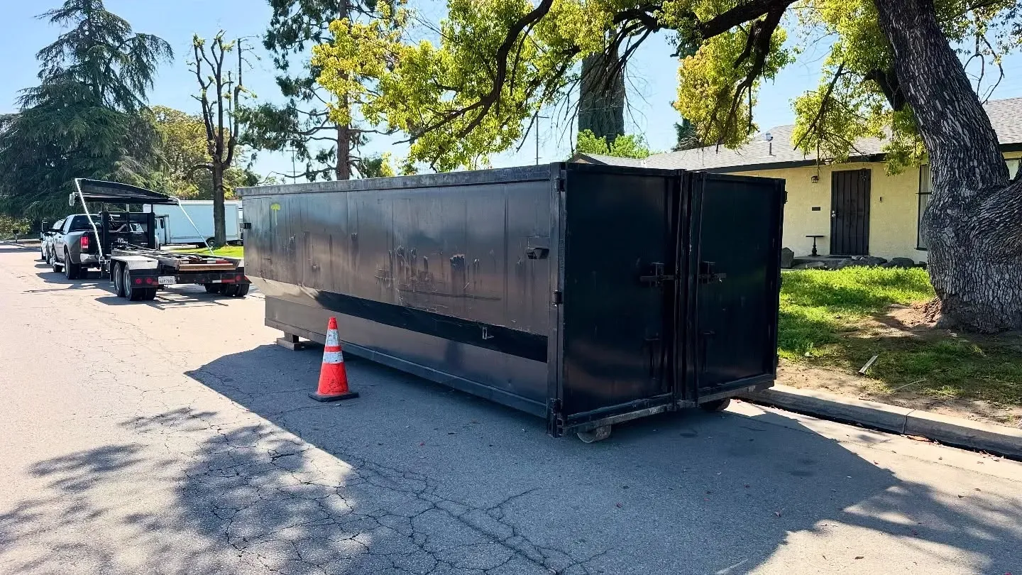 Black dumpster on wheels sits in street next to an orange cone and a tow truck.