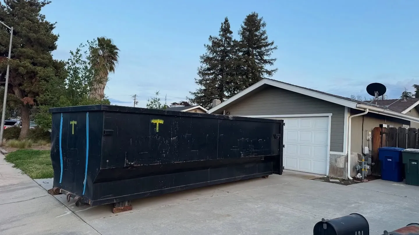 Black dumpster in driveway next to a garage; houses in the background.