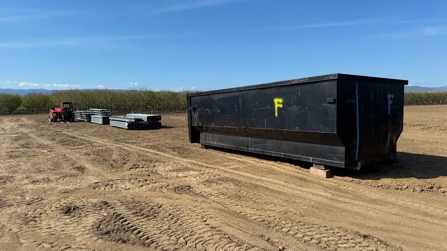Black dumpster in a field, with a tractor and concrete blocks.