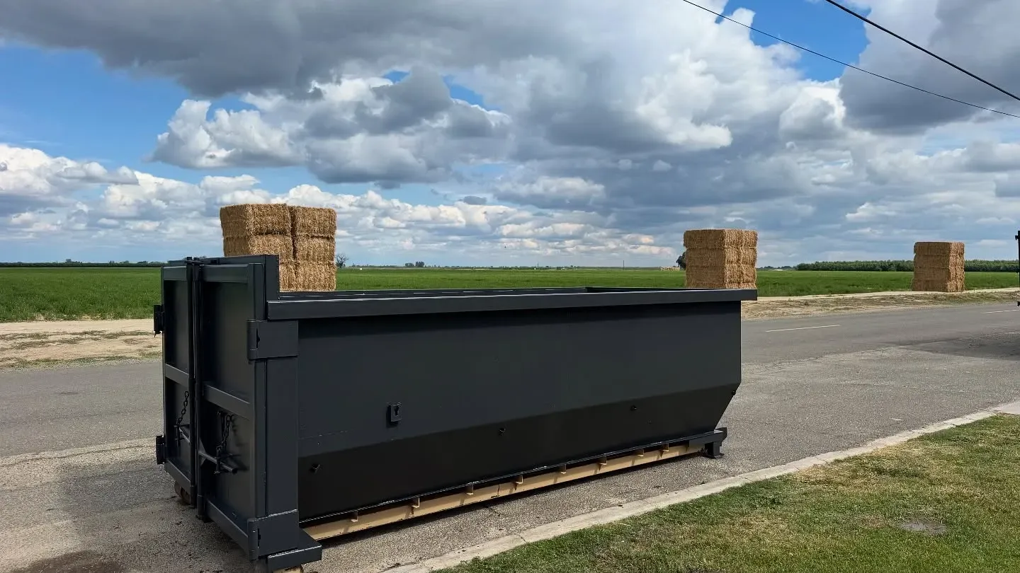 Black dumpster on a paved road with hay bales in a rural setting under a cloudy sky.