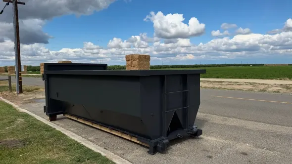 Black dumpster on roadside, with hay bale inside. Field and sky in background.