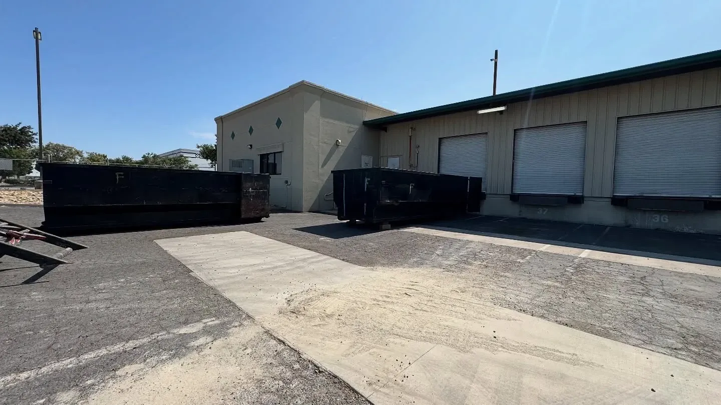 Two black dumpsters sit outside a beige building with loading docks on a sunny day.