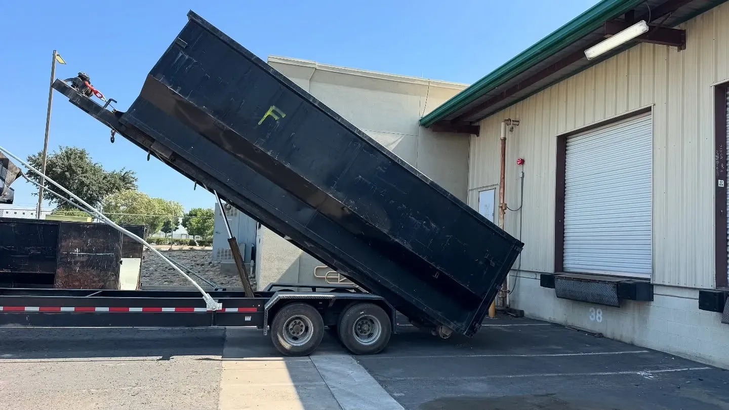 Dump truck unloading near a loading dock; black truck, light building, bright daylight.