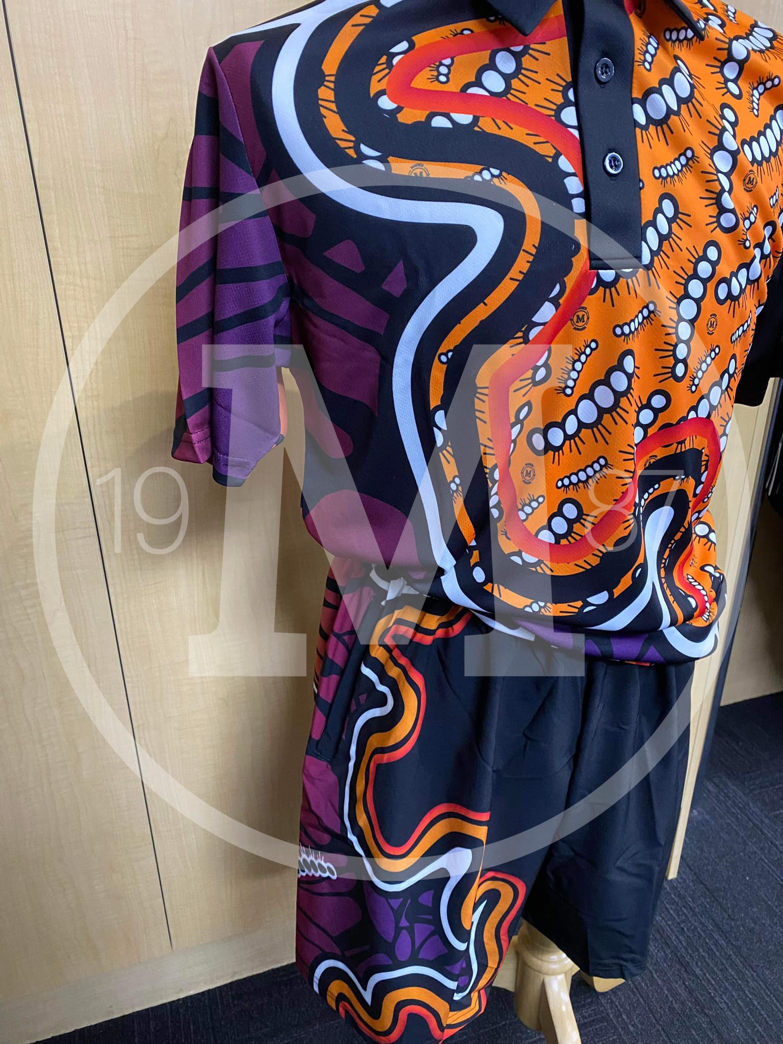 A polo shirt and matching shorts with a vibrant, abstract Indigenous-style pattern in orange, purple, black, and white.