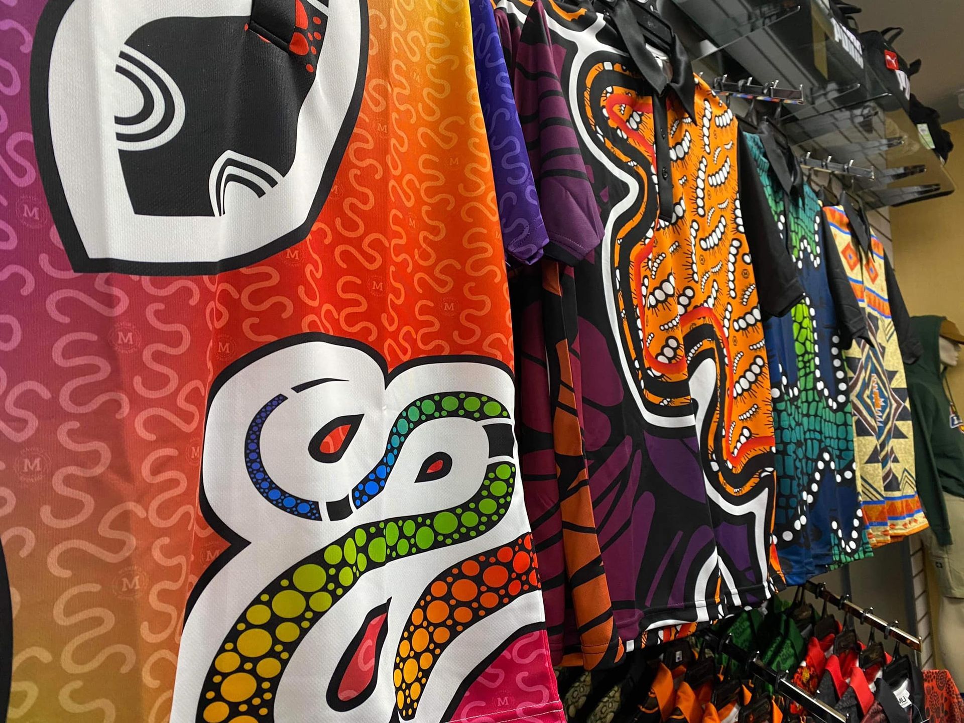 A rack of polo shirts featuring vibrant, colorful Aboriginal-style dot art patterns.