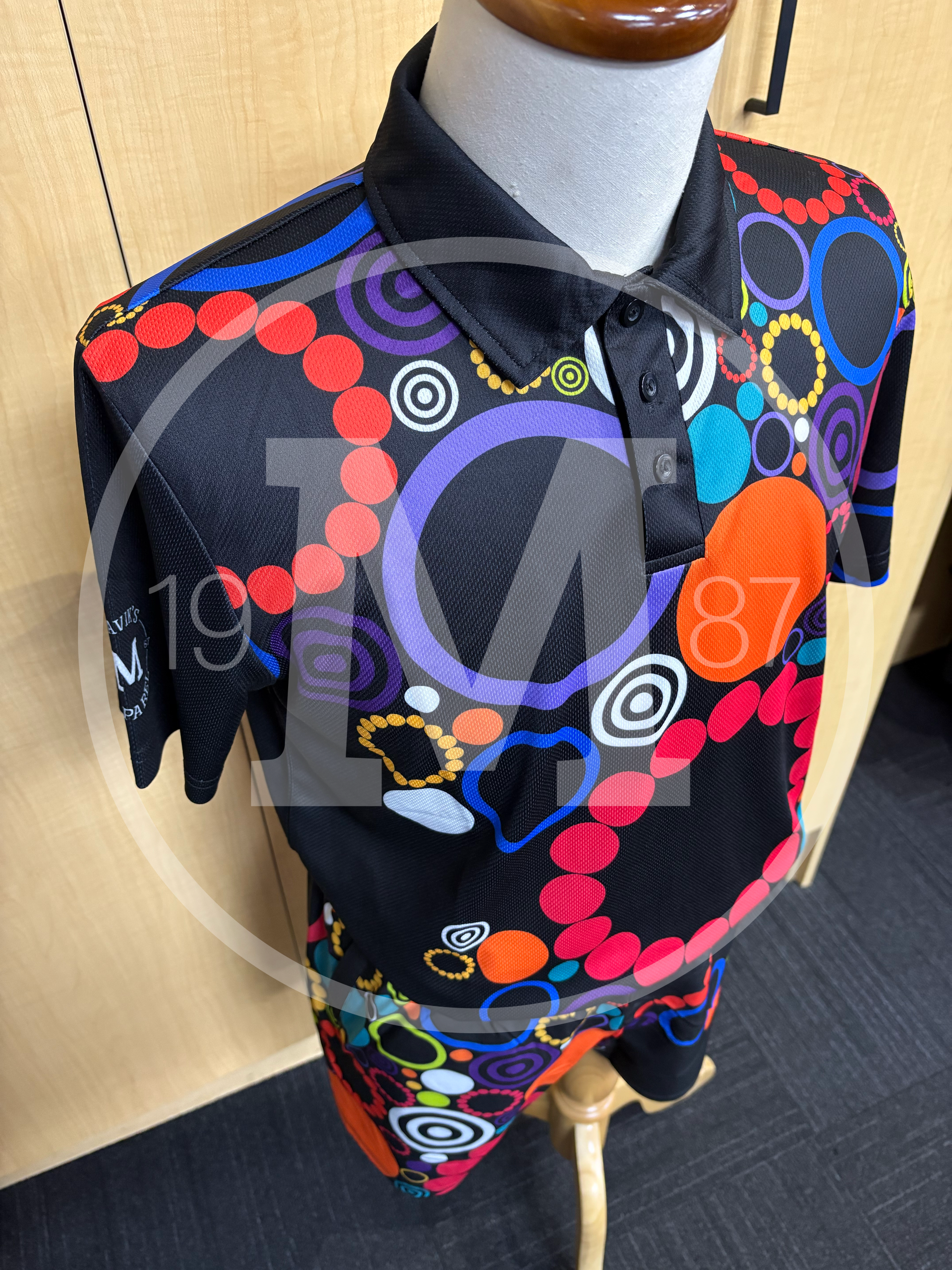 A black polo shirt on a mannequin, covered in a vibrant, colorful pattern of circles, rings, and abstract shapes.