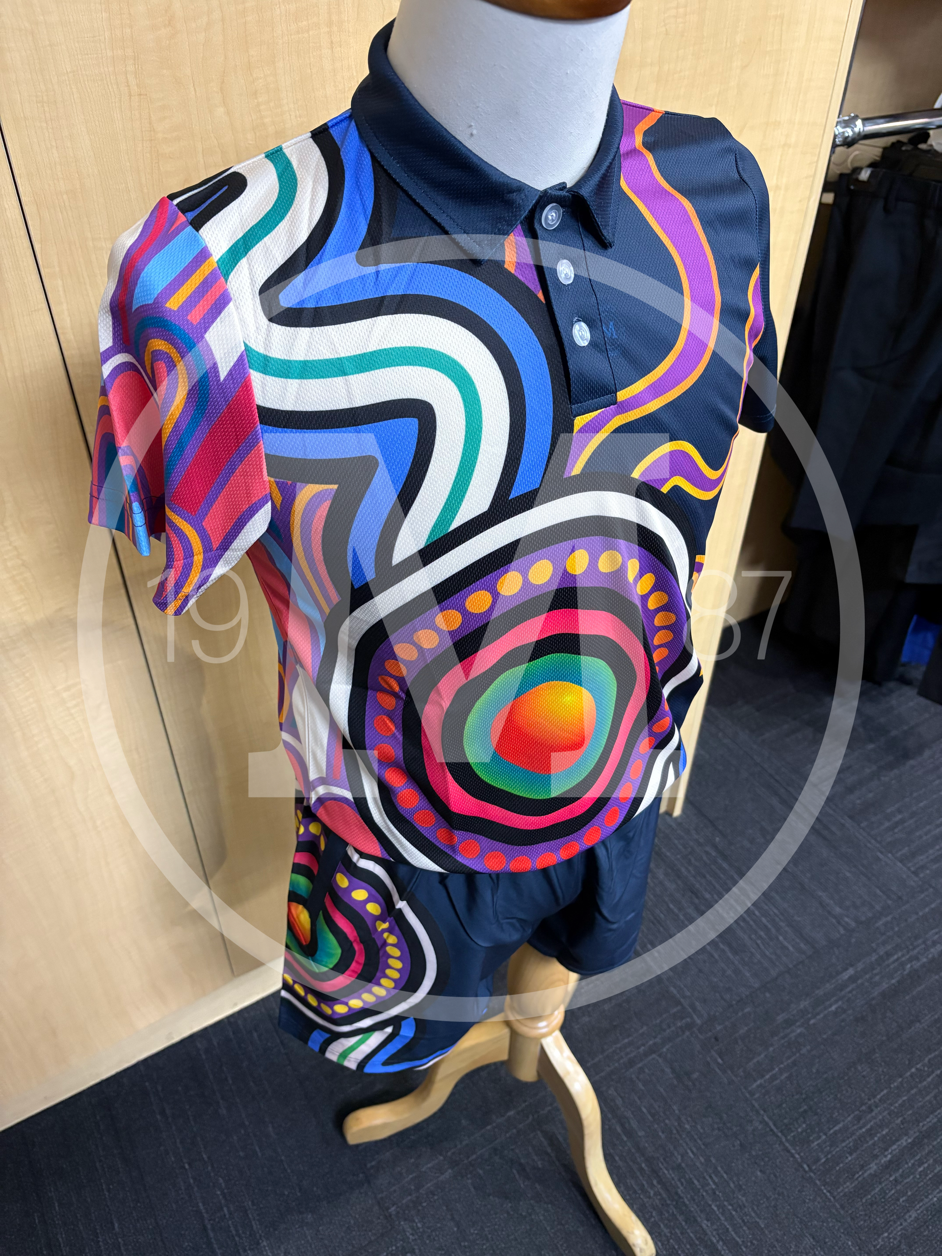 A polo shirt and matching shorts on a mannequin, featuring colorful, abstract Indigenous-style art on a dark blue base.