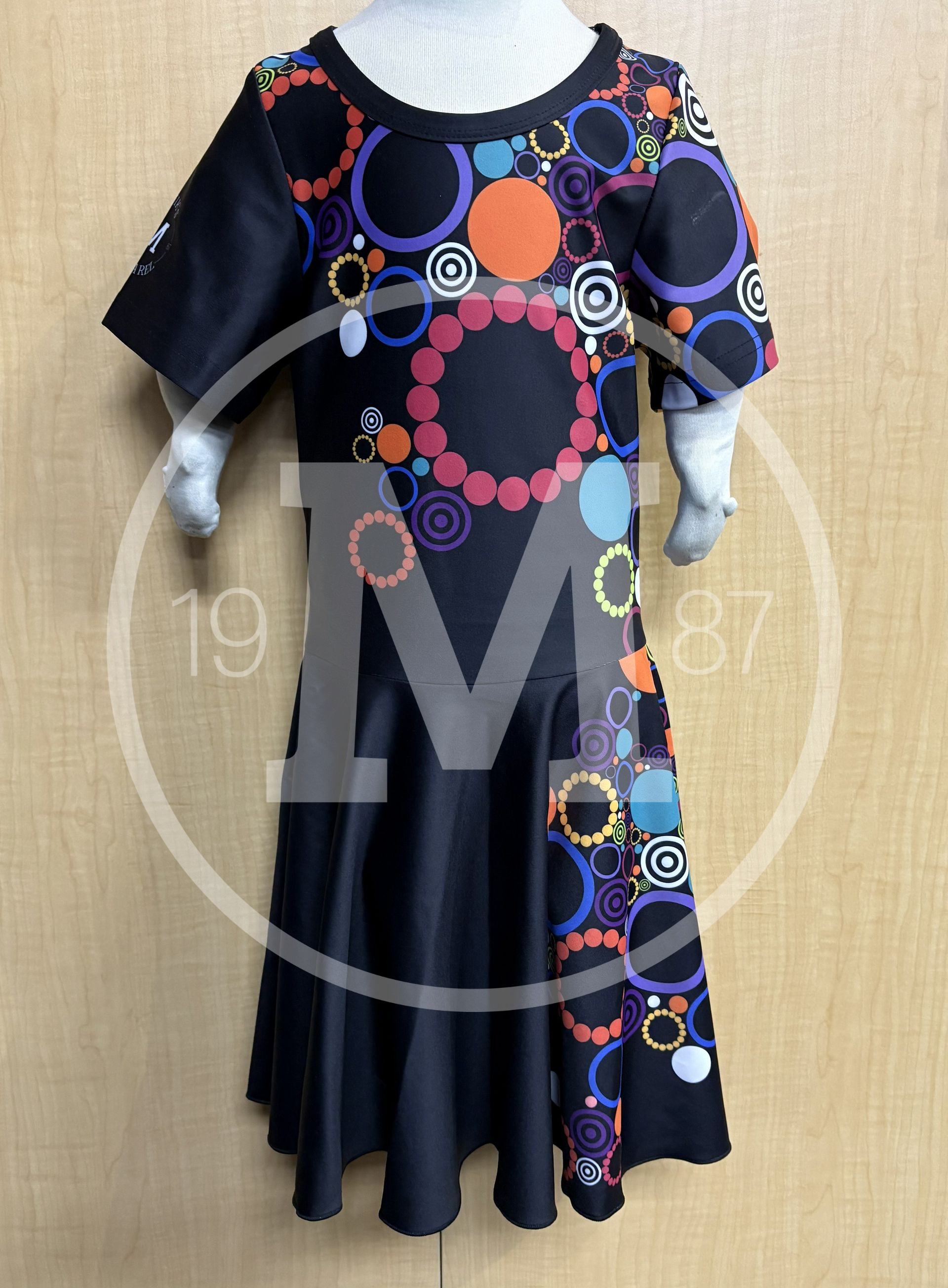 A black dress with a multi-colored circular pattern on the bodice and a flared hem on a mannequin against a wood wall.