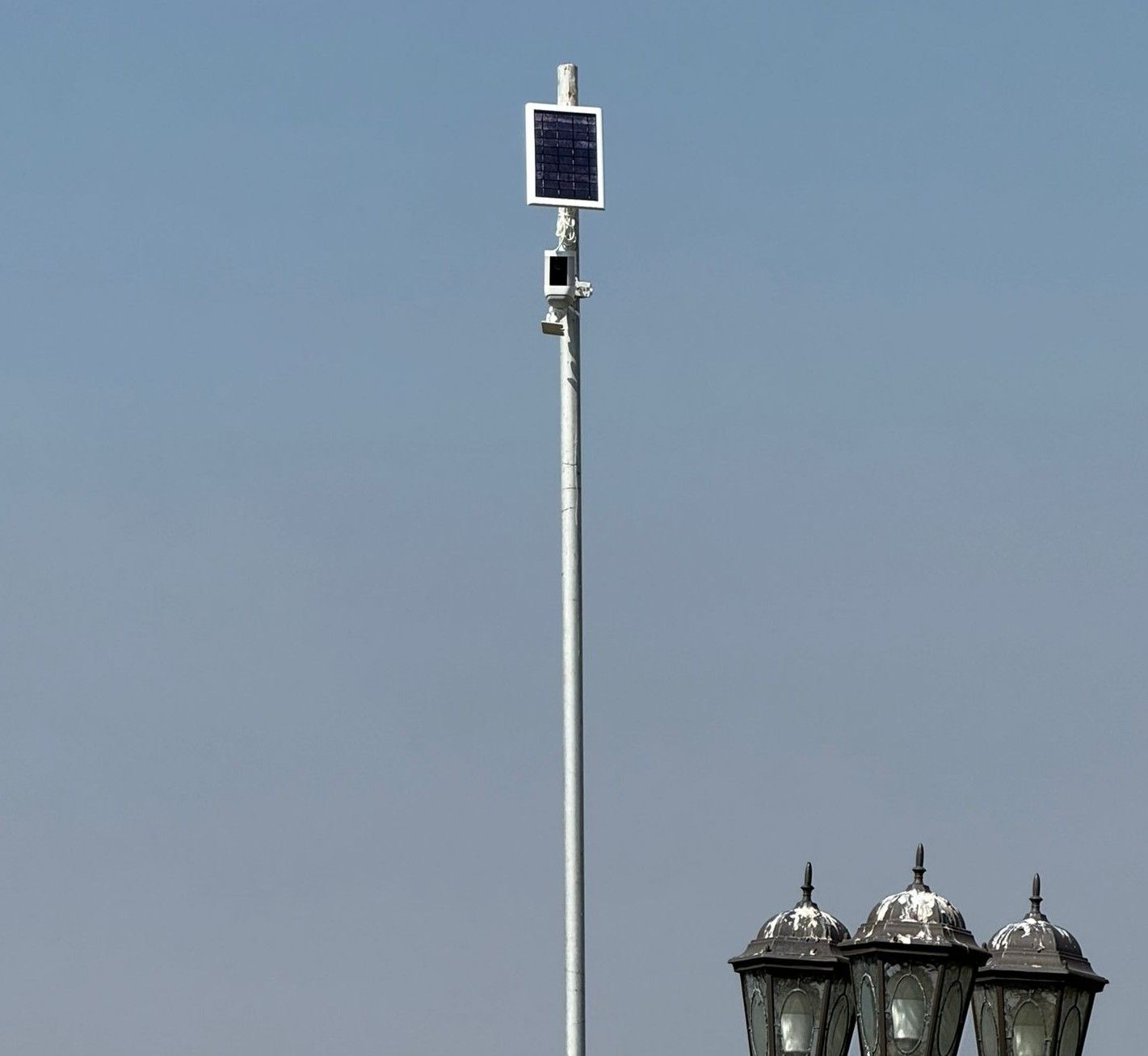 Solar-powered camera on a metal pole, mounted above street lamps against a blue sky.