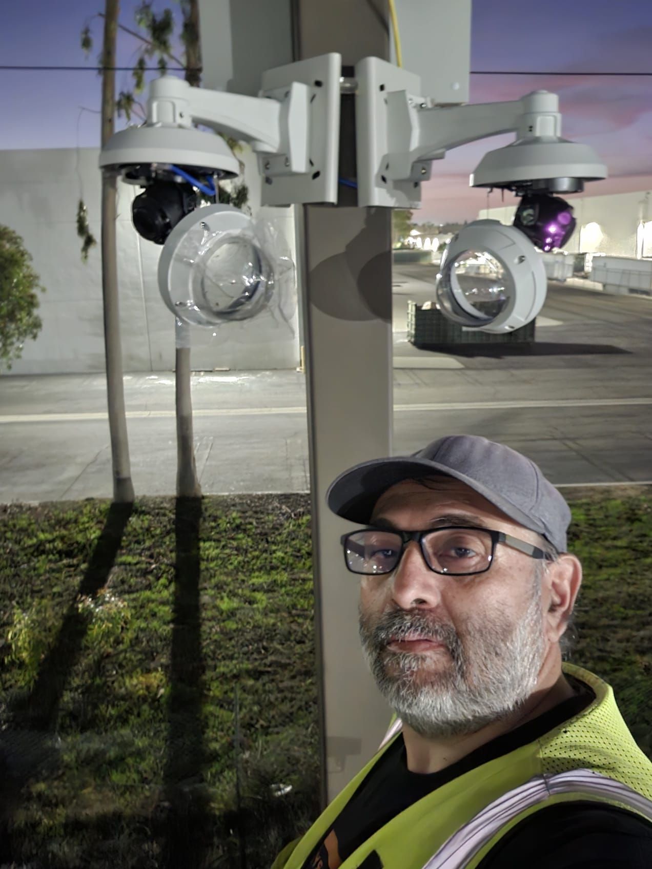 Man in glasses and vest poses near security cameras mounted on a pole outdoors at dusk.