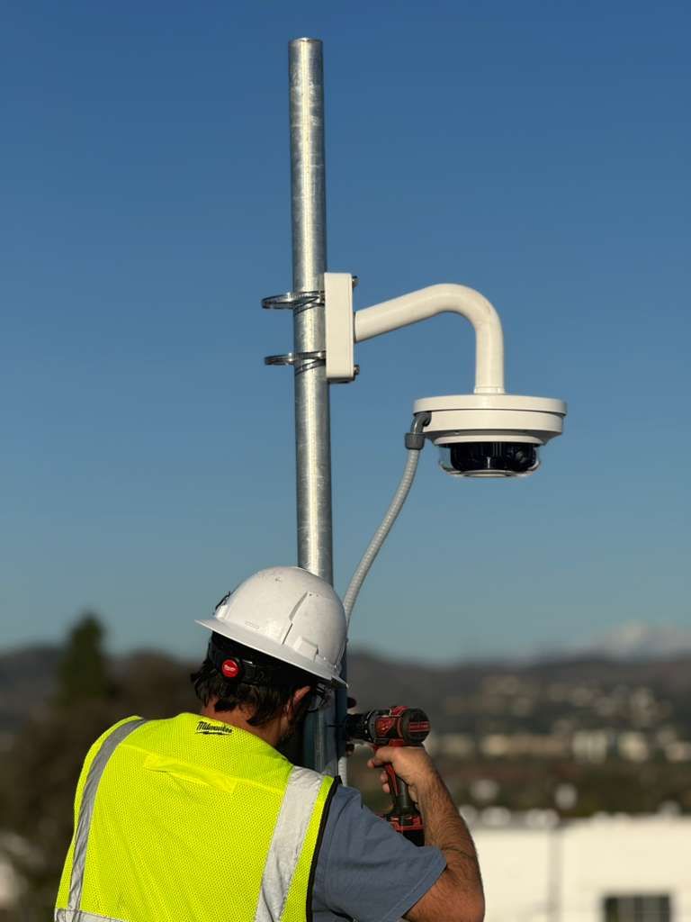 Construction worker installing a security camera on a pole, outdoors under a blue sky.