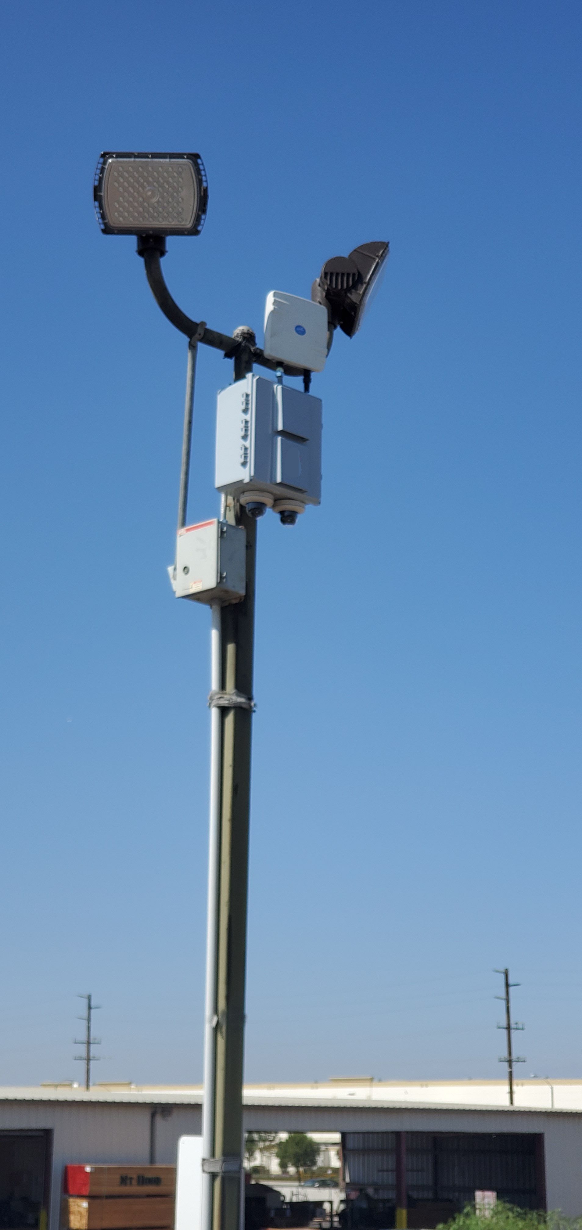 Pole-mounted security equipment with a spotlight, cameras, and electrical boxes against a blue sky.