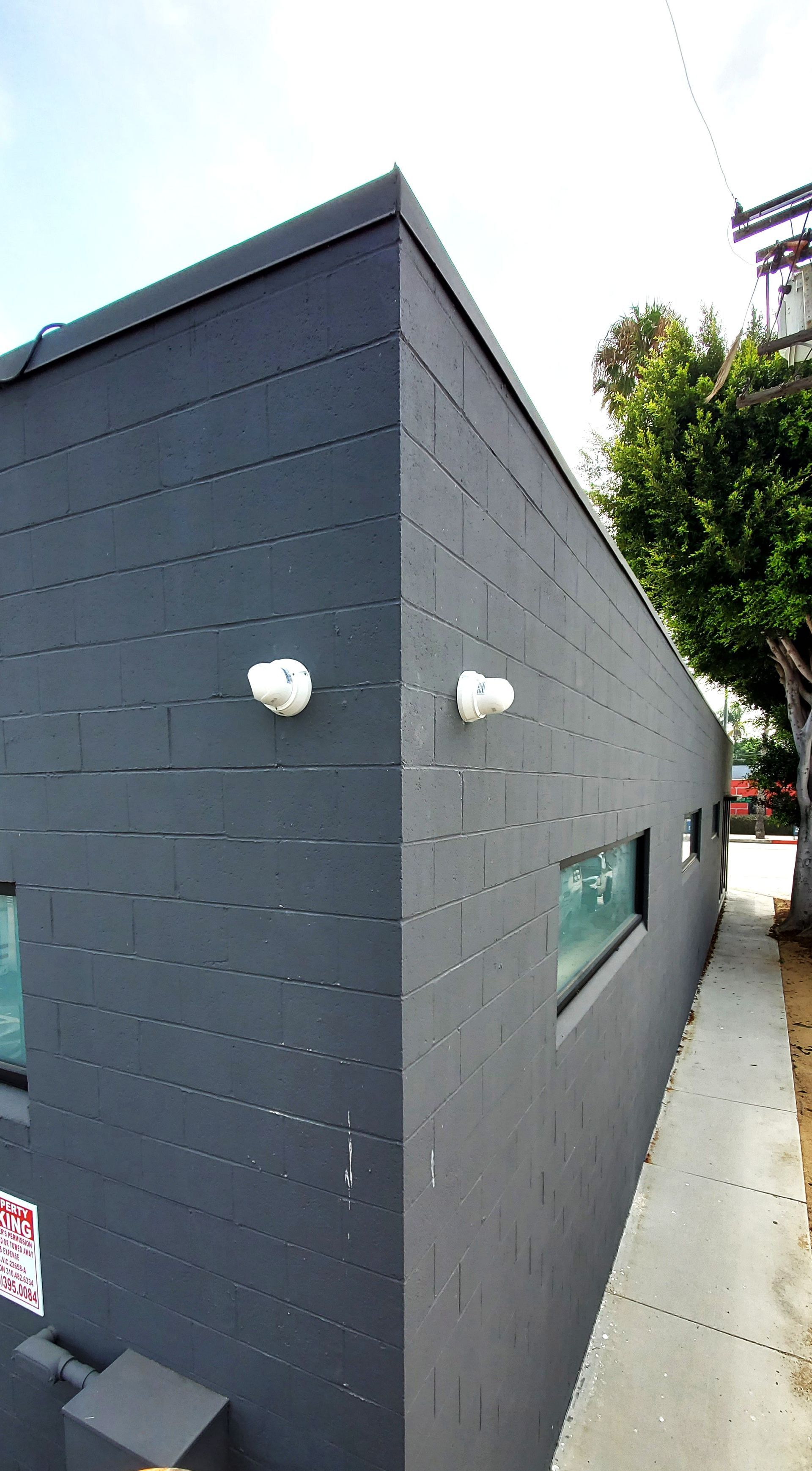 Dark gray building corner with two security cameras and small windows. Sidewalk and trees visible.