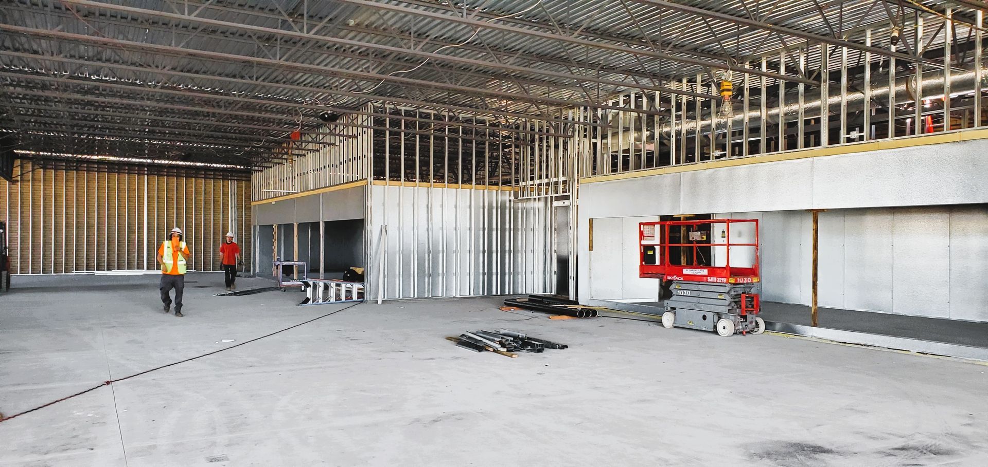 Interior of a building under construction. Two workers in hard hats walk the concrete floor. A scissor lift is on the right.