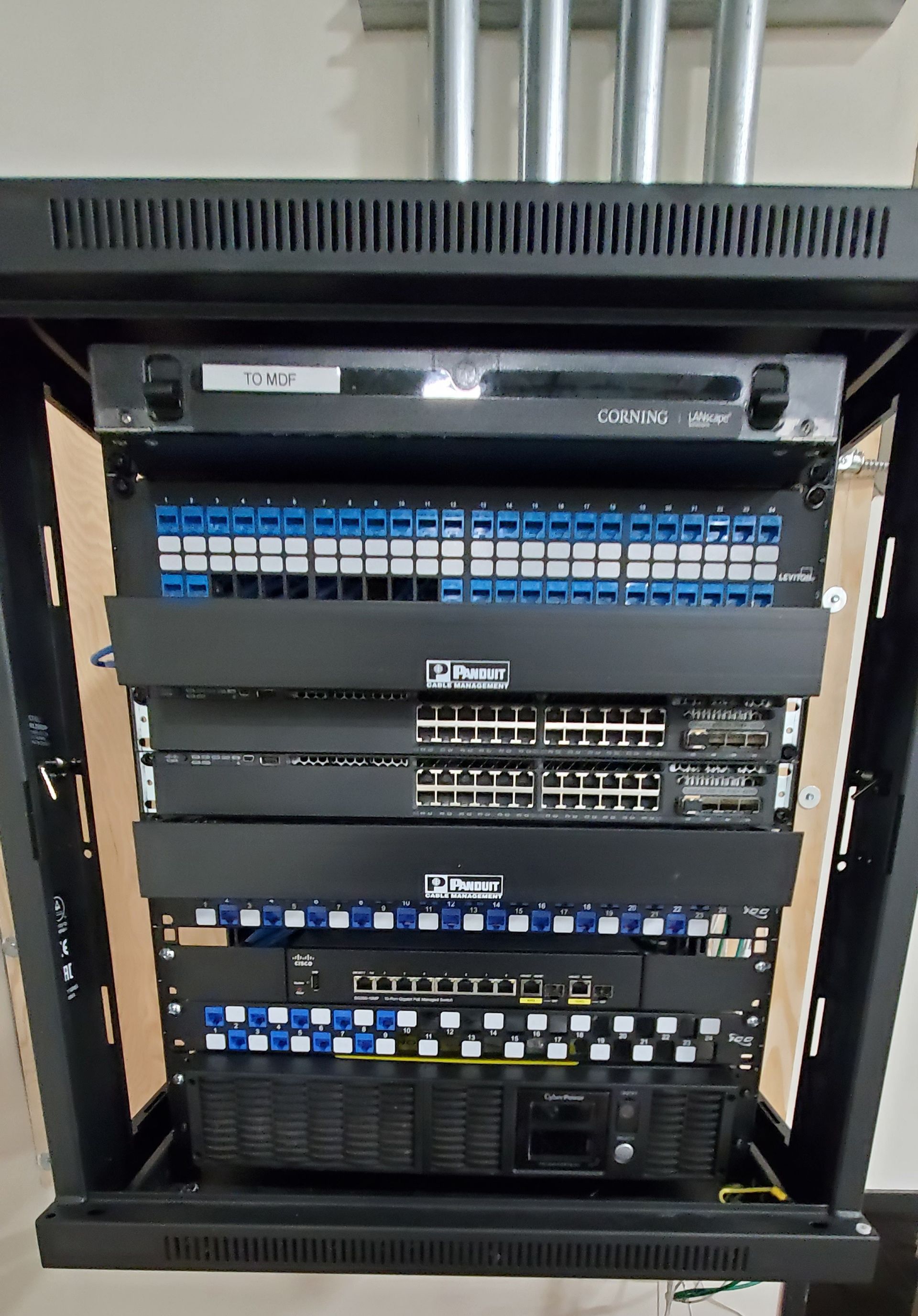 Black network rack with various network devices, cables and patch panels, mounted on a wooden surface.