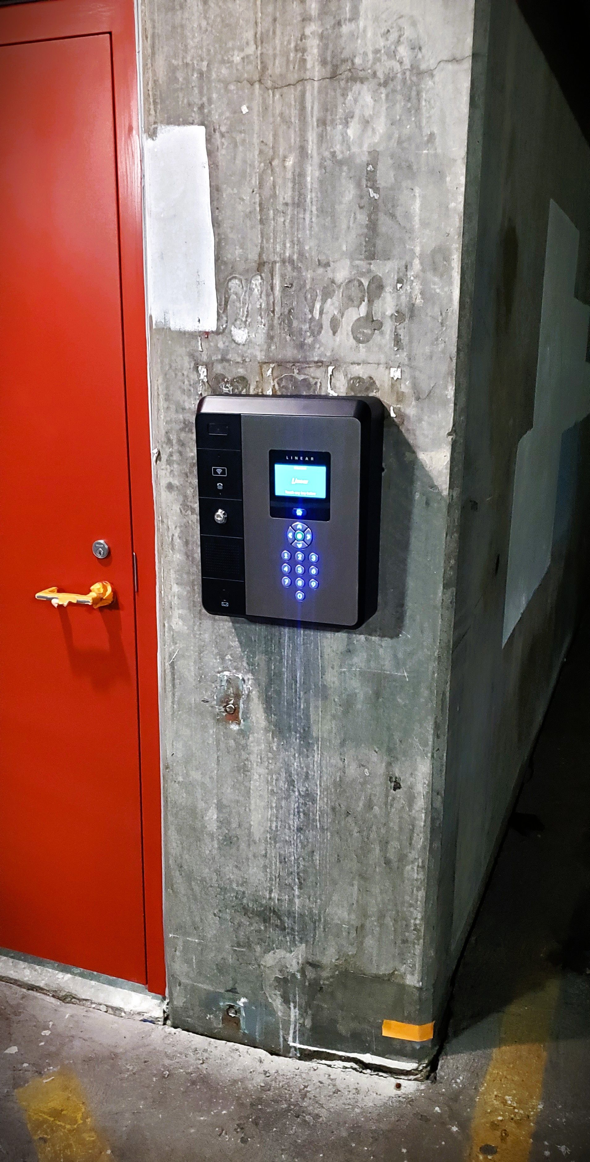 Black access control panel with keypad and screen on concrete pillar near red door.