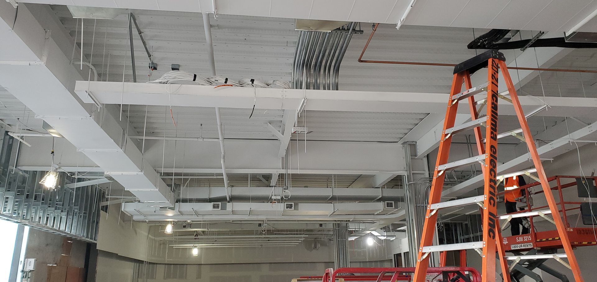 Interior view of a construction site. A tall orange ladder rests against a white ceiling, with exposed wiring and ductwork.