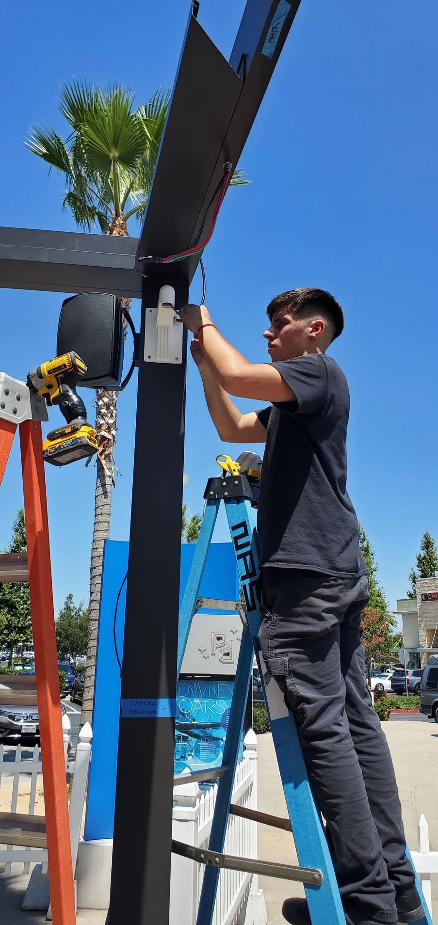 Person on a ladder installing something on a black beam outdoors on a sunny day.