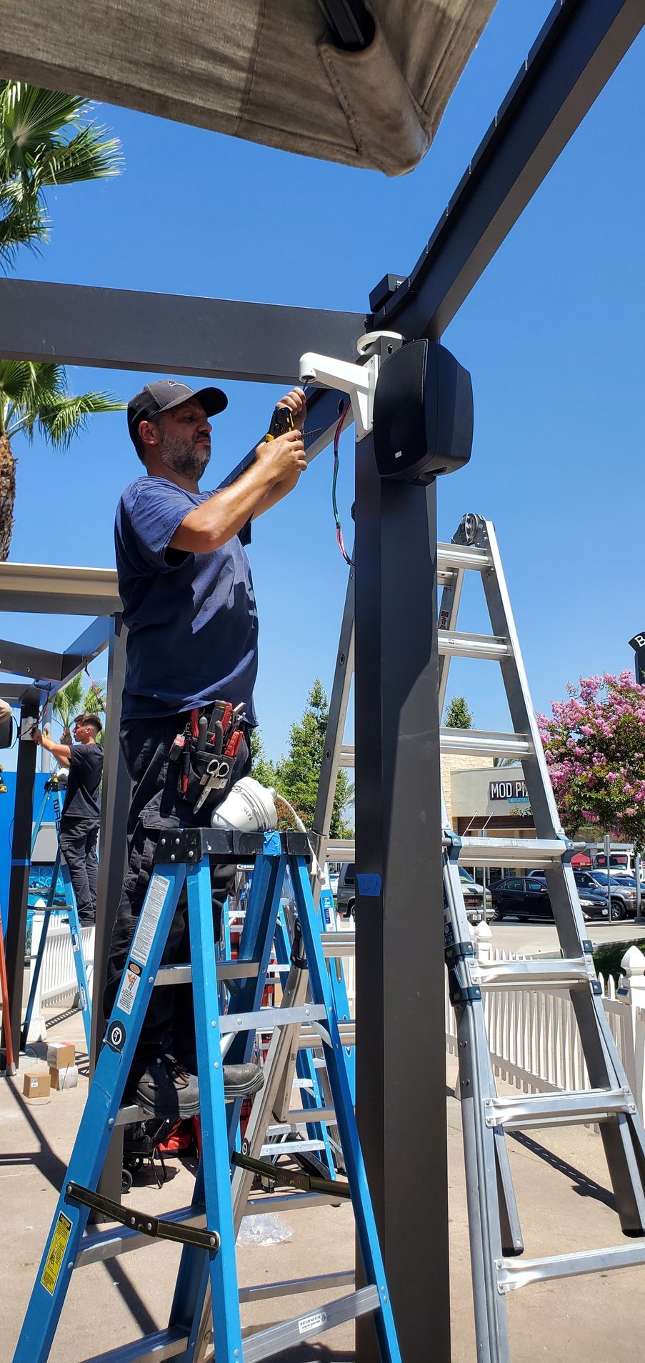Man on ladder installing awning on a sunny day. Two ladders and another person are also present.