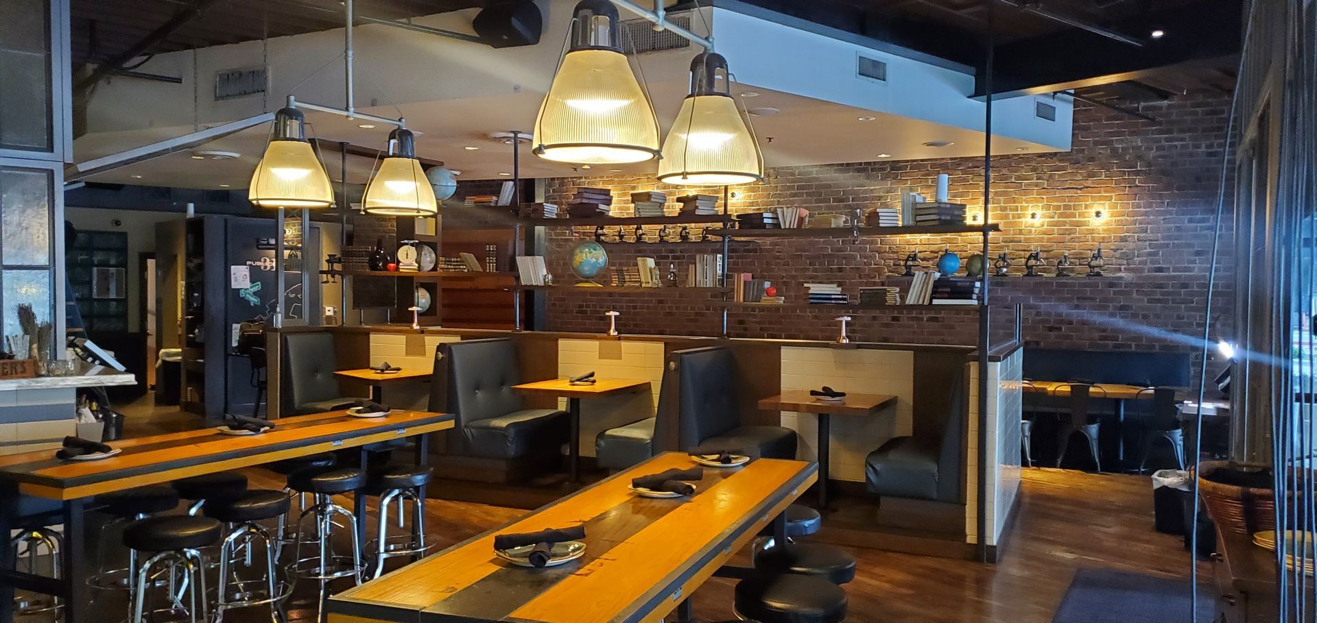 Interior view of a restaurant with wooden tables, booths, and pendant lights. A brick wall with shelves is visible.