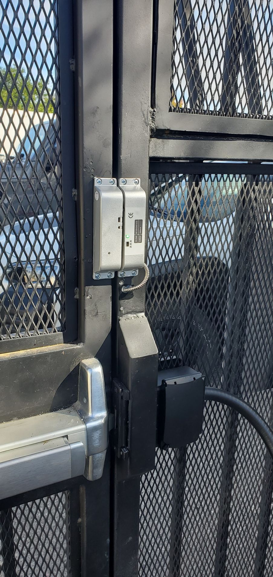 Close-up of a black metal gate with a latch, electrical box, and a push bar for opening.
