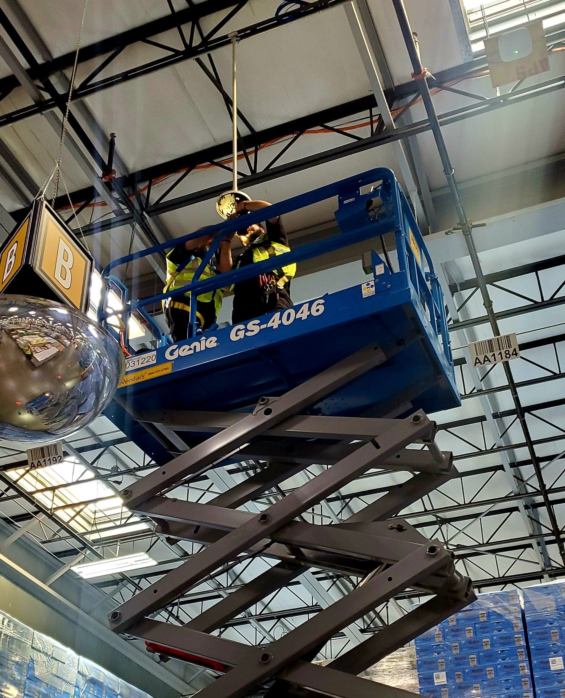 Two workers on a blue scissor lift inside a building, working on overhead fixtures.