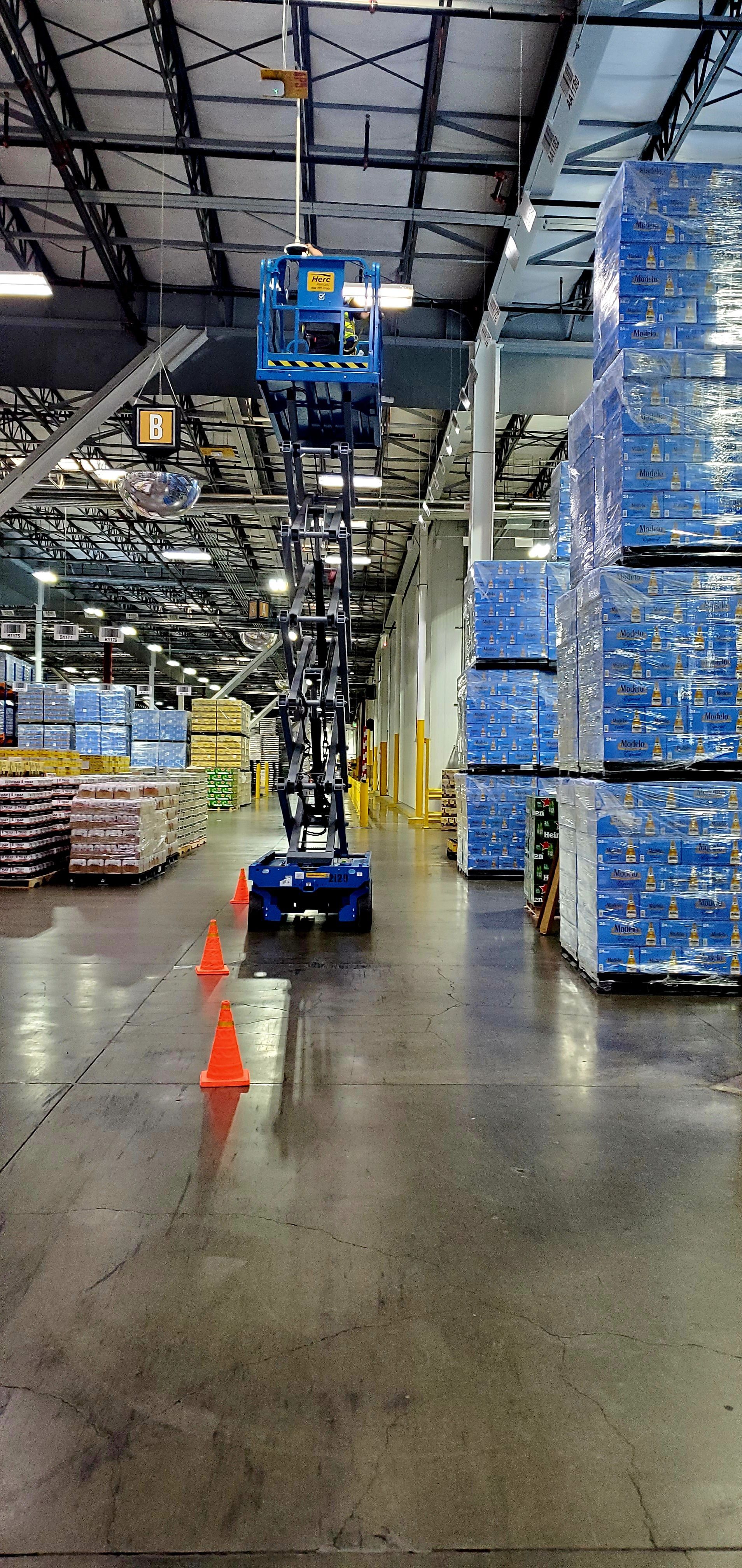 A blue lift in a warehouse with orange cones on the floor. Pallets of packaged goods are stacked.