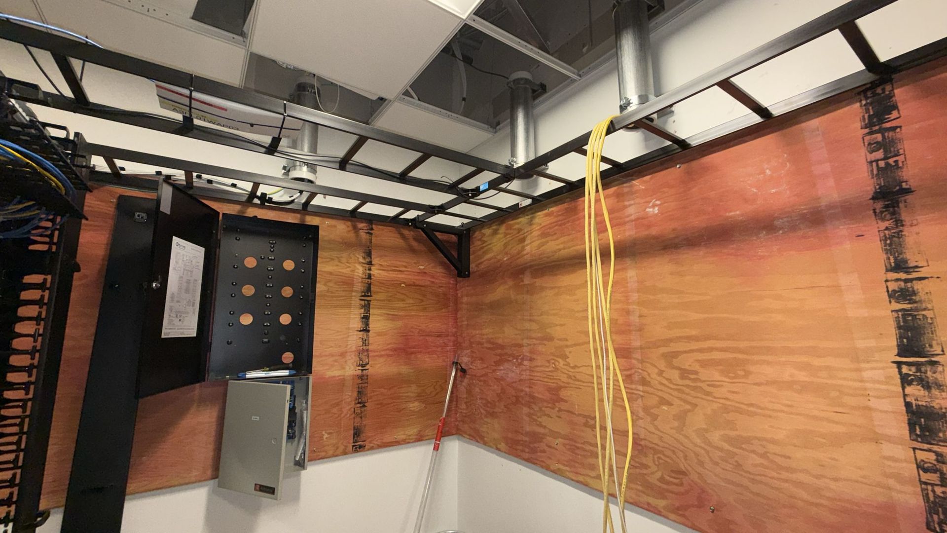 Wooden-walled room with electrical equipment, cables, and yellow cord hanging from the ceiling.