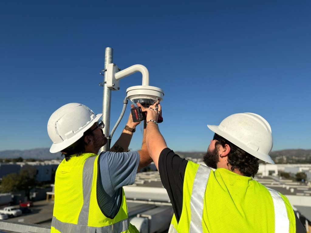 Two workers in hard hats and vests install a security camera on a rooftop under a blue sky.