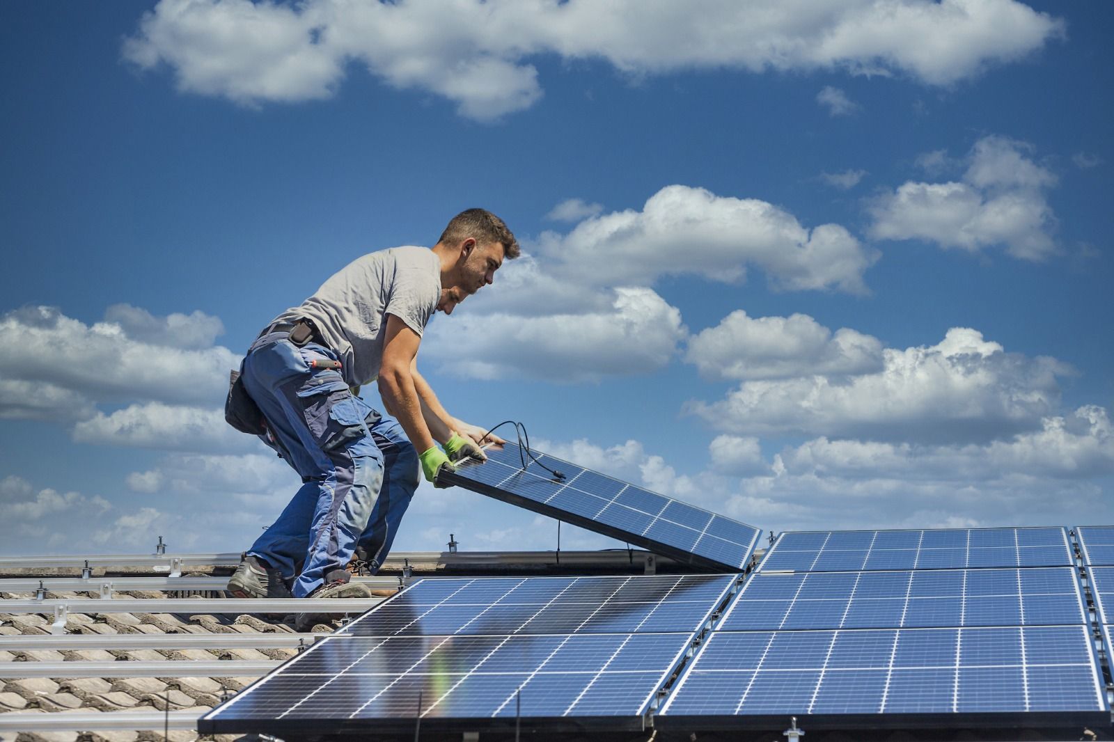 Worker installing solar panel on a rooftop under a cloudy blue sky.
