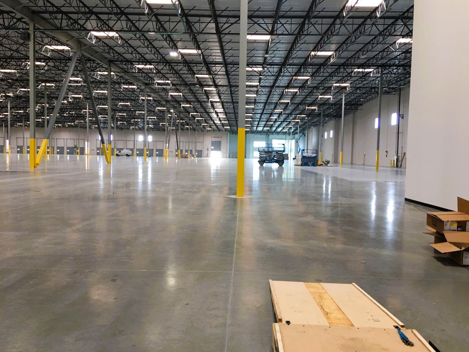 Large, empty warehouse interior. Concrete floor, high ceilings, bright lighting. Black beams, yellow support poles.