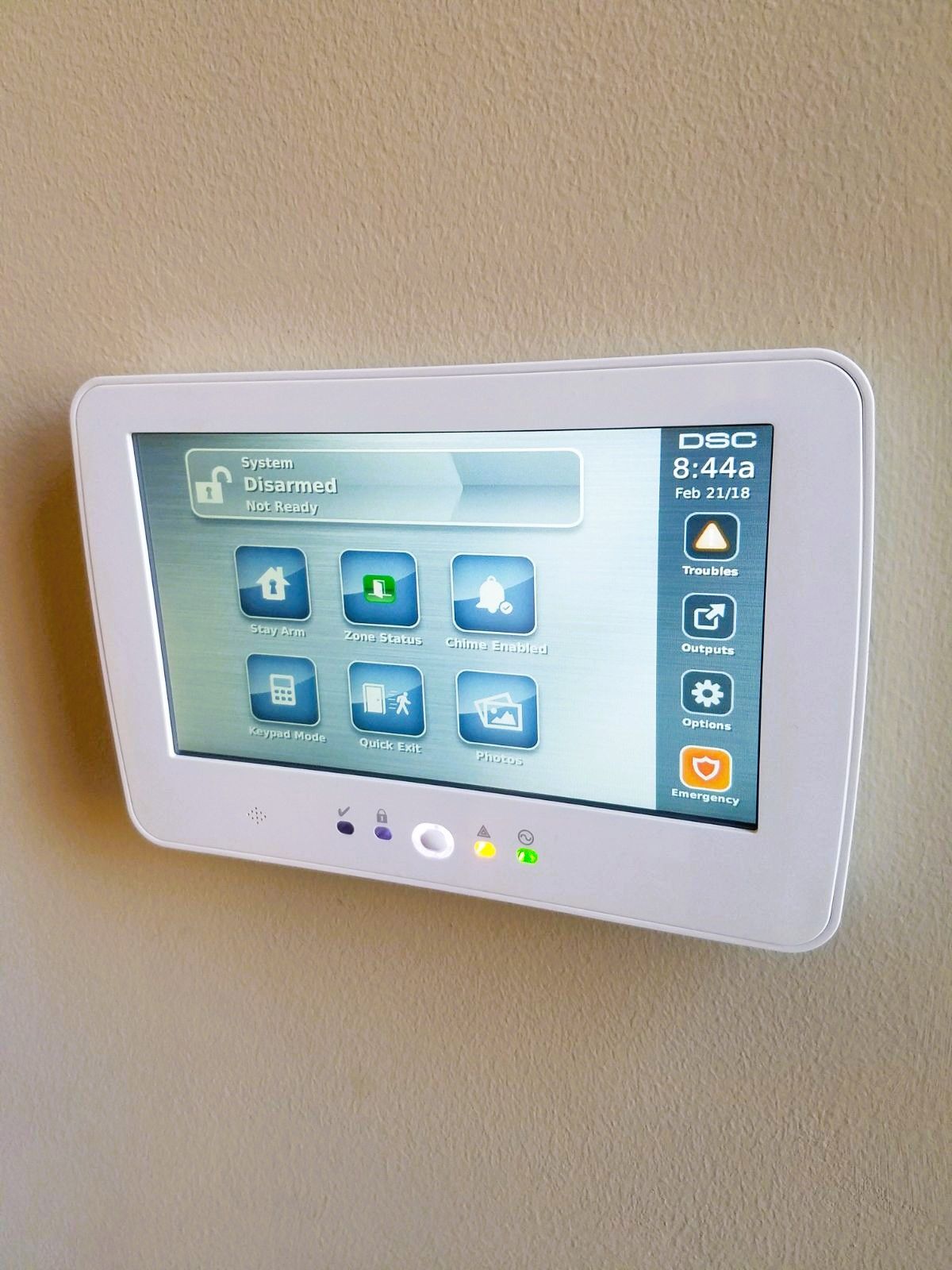 White security system control panel mounted on a beige wall, displaying various icons and the time.