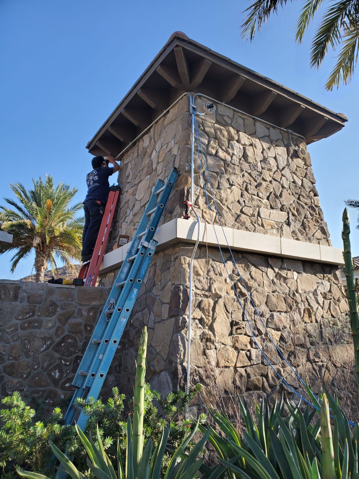 Person on ladder installing electrical wiring on stone building under blue sky.
