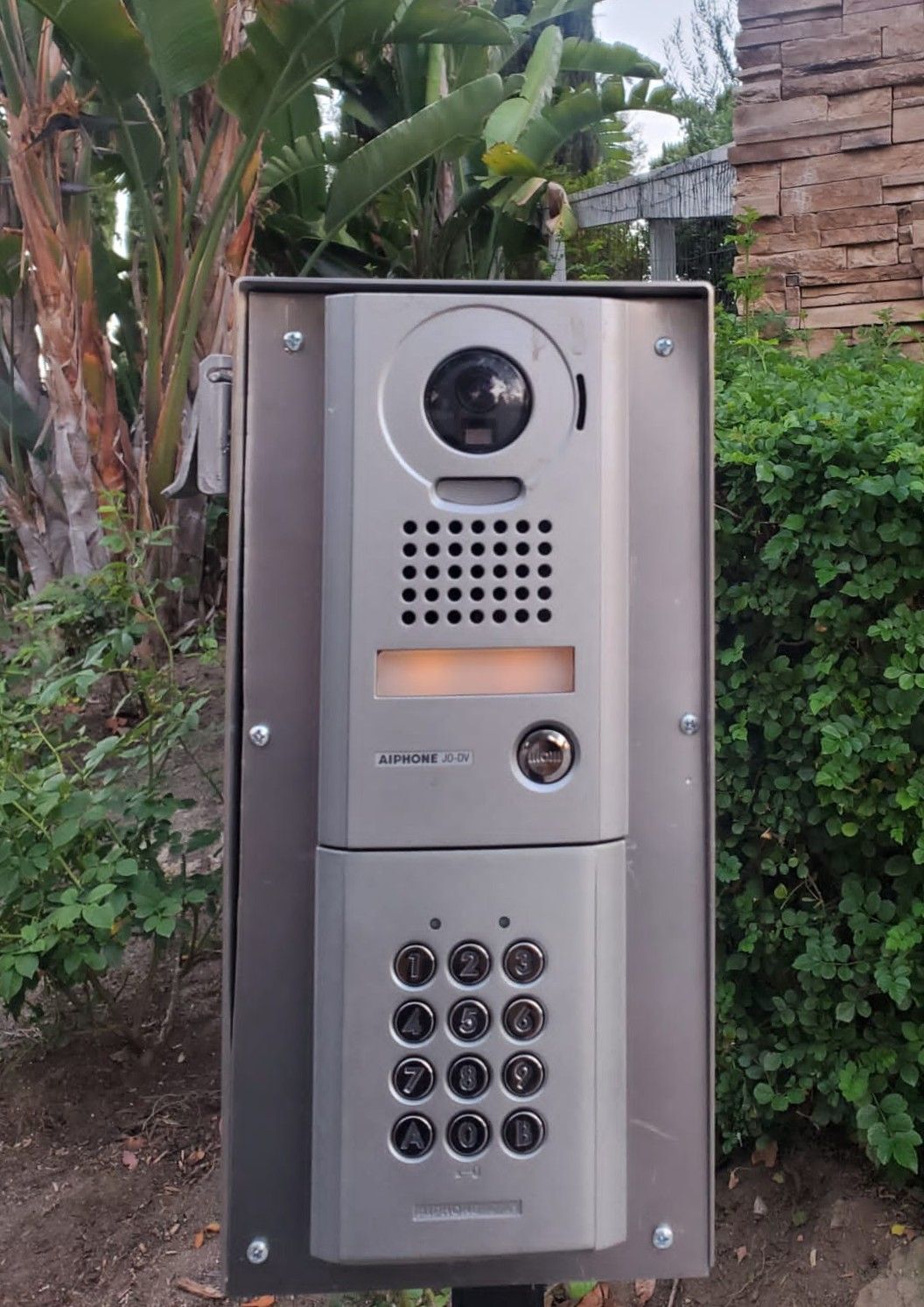 Silver intercom system with camera, keypad, and speaker mounted on a post, outdoors.