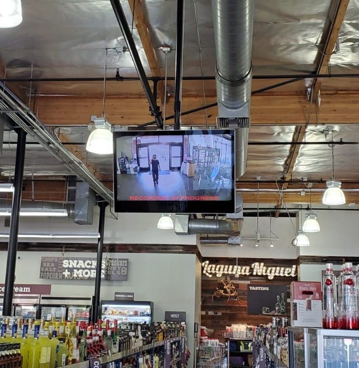 TV screen in a liquor store showing security footage. Store interior with liquor bottles, ceiling beams, and lights.