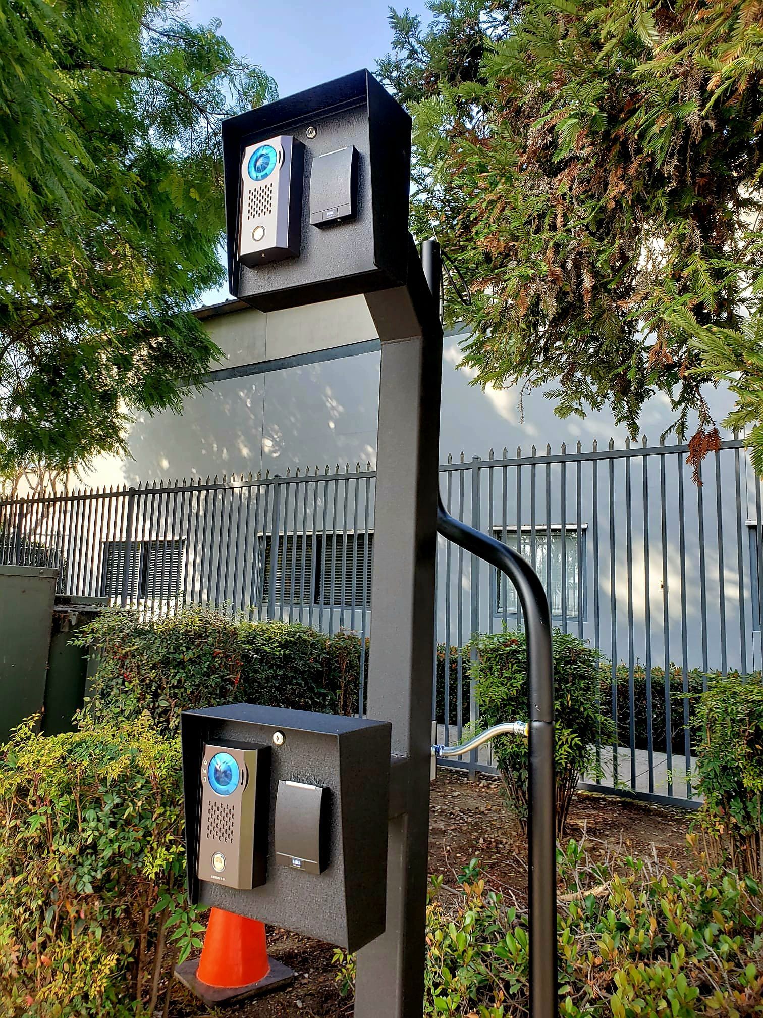 Black intercom system on pole with two call boxes, an orange cone, and a chain-link fence in the background.
