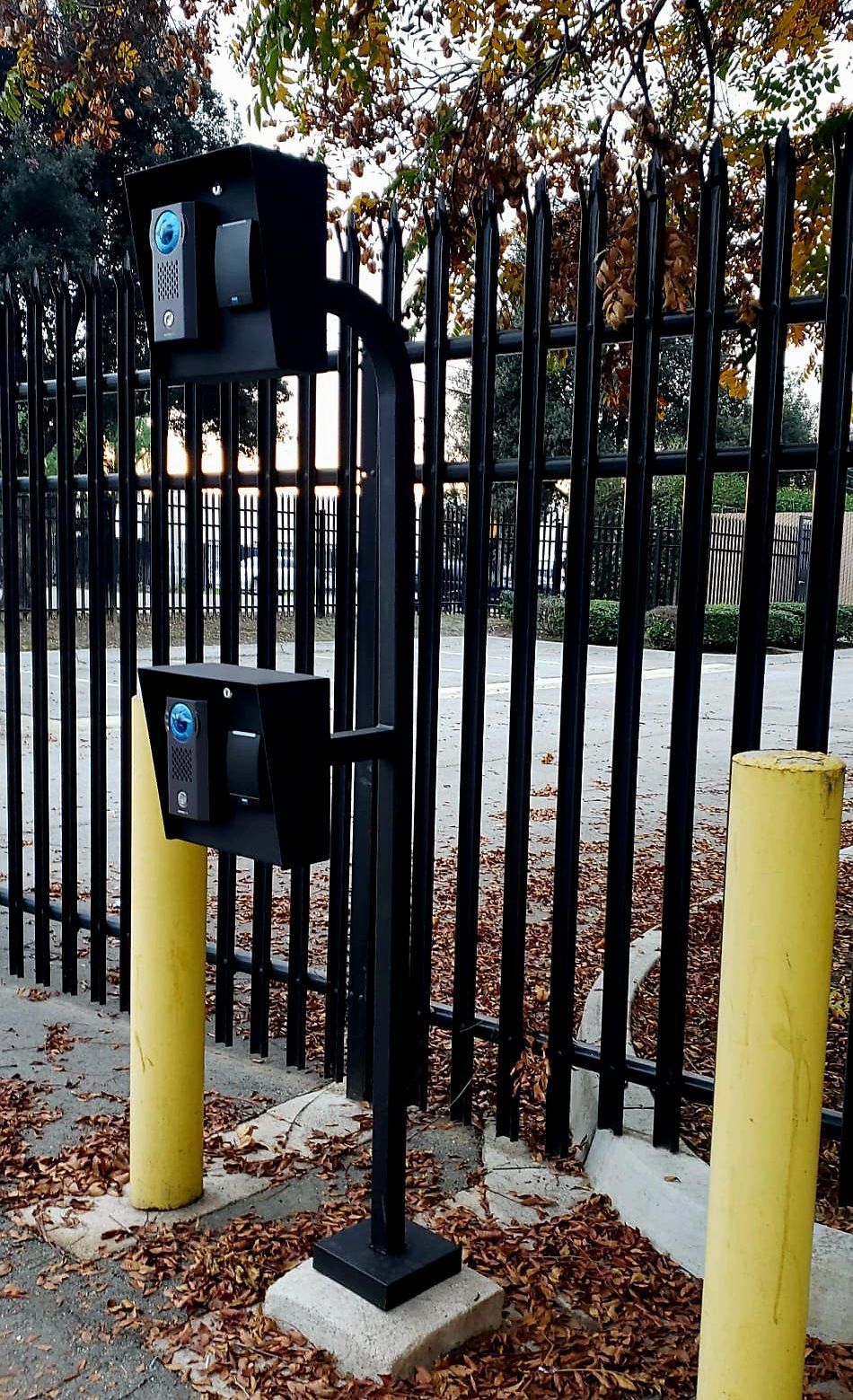 Black gate access control devices mounted on a pole with a black fence in the background. Two yellow bollards flank the pole.