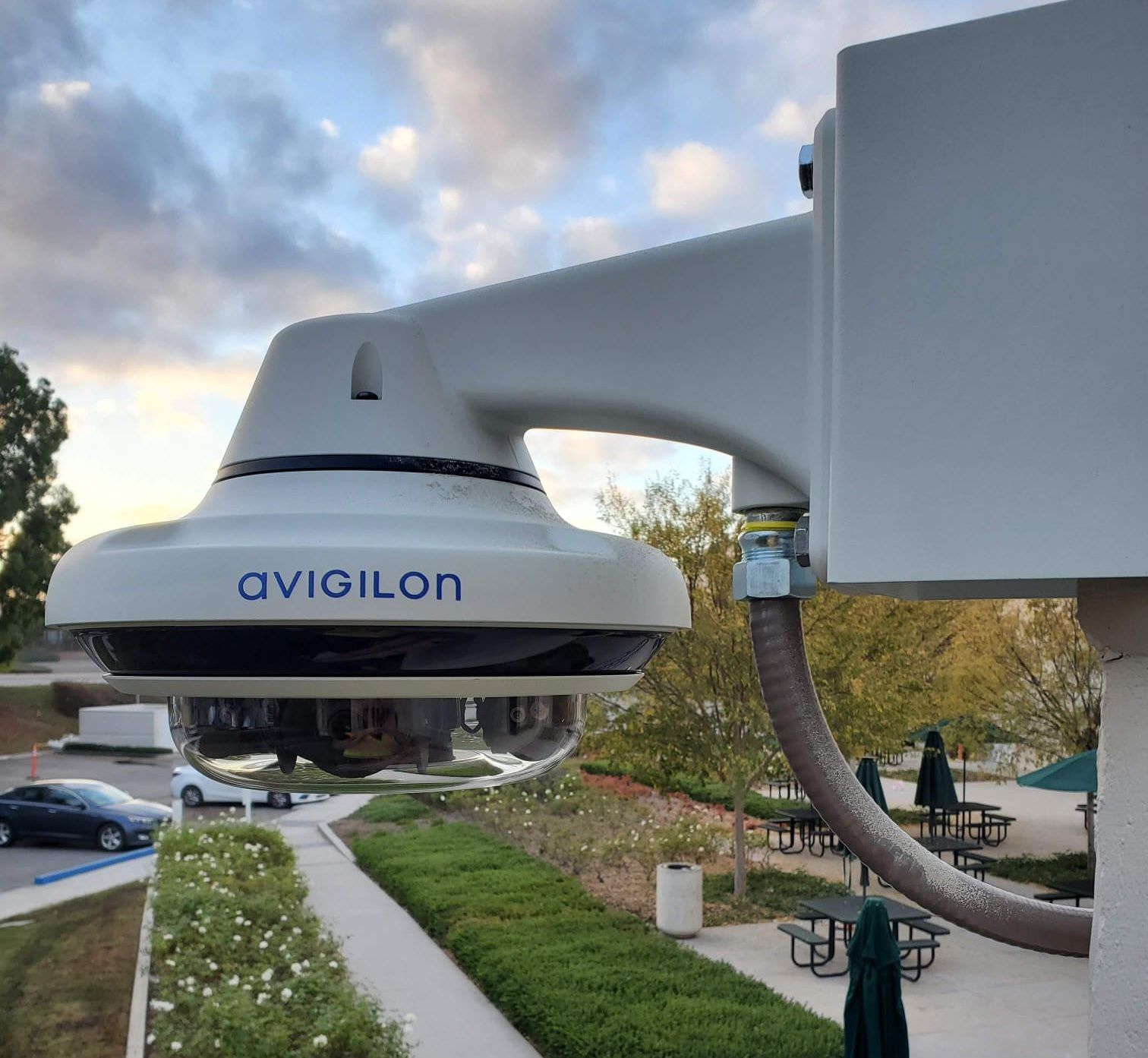Security camera mounted on a building, overlooking an outdoor area with greenery and cars.