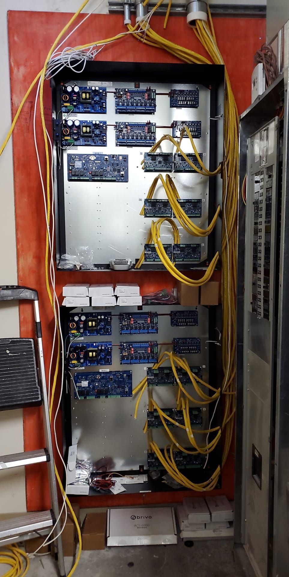Two computer server units mounted on a red wall, with circuit boards and yellow cabling. Ladder and storage nearby.