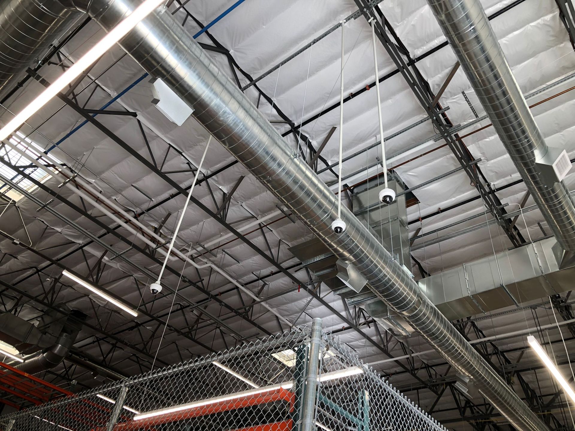 View from below of a high ceiling with metal beams, ductwork, security cameras, and fluorescent lights.